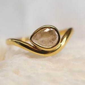 May include: A gold ring featuring a pear-shaped, faceted, light-colored stone set in a smooth bezel. The band has a curved, wave-like design that tapers toward the setting, creating a continuous, polished metallic surface around the central gemstone.