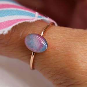 May include: A thin gold-toned metal ring featuring a small, smooth, oval-shaped cabochon stone. The stone displays a blended pattern of soft pink, light blue, and white colors, resembling a watercolor effect. The ring band is simple and polished.