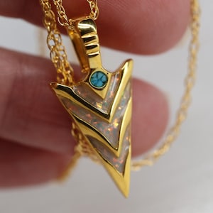 May include: Gold-colored pendant necklace featuring an arrowhead design. The arrowhead has a turquoise accent and is inlaid with white and iridescent material. The necklace chain is also gold-colored.
