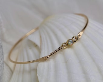 May include: A thin gold bangle bracelet featuring a single small round bezel-set opal gemstone at the center. The bracelet has a hook-and-eye style closure mechanism integrated into the design, showcasing a polished metallic finish and iridescent stone.