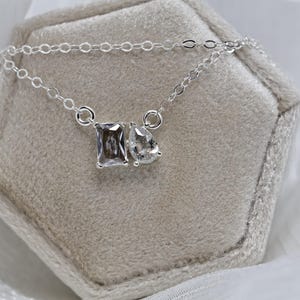 May include: A silver necklace with two clear gemstones, one rectangular and one pear-shaped, displayed on a gray velvet jewelry box. The necklace chain is delicate and the gemstones sparkle.