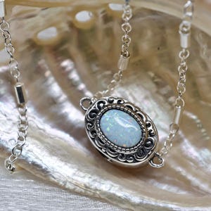 May include: A silver chain necklace with a white opal pendant set in a silver filigree frame. The necklace is displayed on a white iridescent shell.