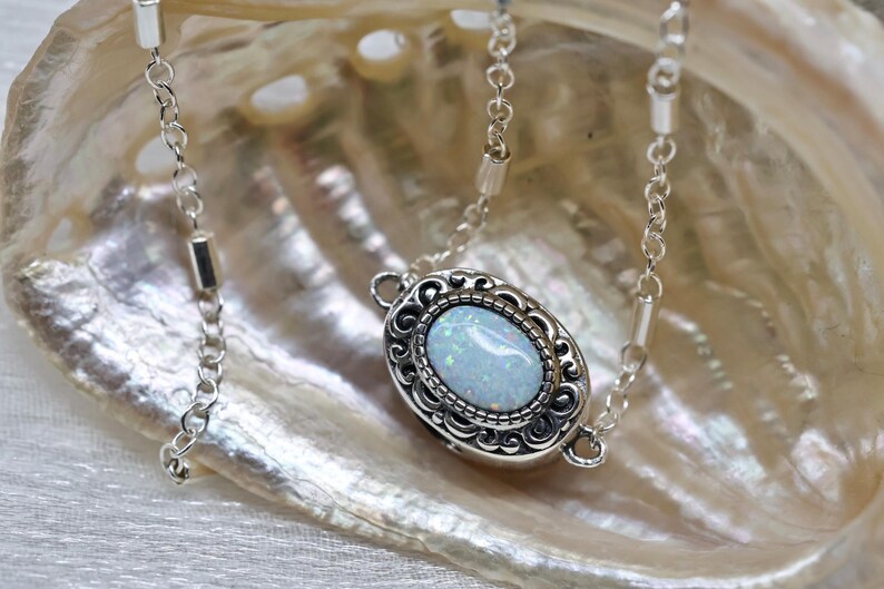 May include: A silver chain necklace with a white opal pendant set in a silver filigree frame. The necklace is displayed on a white iridescent shell.