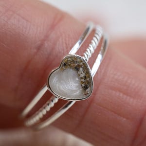 May include: A silver ring featuring a heart-shaped setting. The heart contains a white, pearlescent inlay on one side and a textured, sandy-colored granular material on the other. The band consists of two thin, smooth silver wires and one twisted wire.