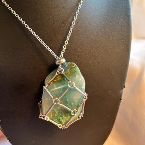 Puede incluir: Collar de cadena plateada con un colgante de piedra verde y dorada de forma irregular. La piedra está encerrada en una jaula de alambre plateado. El collar se exhibe sobre un fondo oscuro.