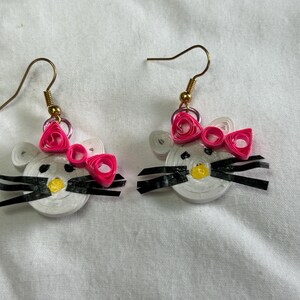 Puede incluir: Pendientes hechos a mano con un diseño de cara de gato en papel quilling blanco, un lazo rosa, bigotes negros y una nariz amarilla. Los pendientes tienen ganchos para las orejas de color dorado.