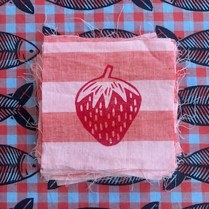 May include: A stack of square fabric swatches featuring a red strawberry print on a light pink and white horizontal striped background. The swatches have raw, frayed edges and are layered on a red and blue checkered fabric with black fish motifs.