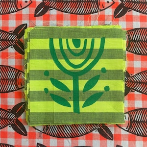 May include: A stack of square fabric swatches featuring a green and lime green horizontal striped pattern with a dark green stylized flower print in the center, layered over a red and white checkered fabric with a black fish skeleton pattern.
