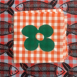 May include: A stack of square fabric swatches featuring an orange and white gingham pattern with a centered dark green four-petal flower print, layered over a background fabric with a red and white gingham pattern and black fish skeleton motifs.