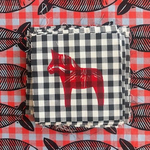 May include: A stack of square fabric pieces featuring a black and white gingham pattern with a red Dala horse print centered on the top piece. The stack rests on a red and white checkered fabric background printed with black fish skeleton motifs.