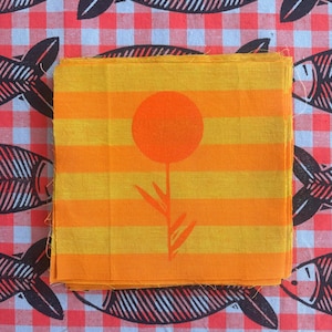 May include: A stack of square fabric swatches featuring horizontal yellow and orange stripes with a centered orange circular flower graphic. The fabric is placed on a red and white checkered background patterned with black fish skeletons.