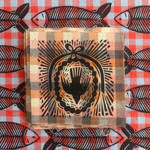 May include: A stack of square fabric swatches featuring a black block-printed bird inside an oval frame with radiating lines, set against a tan and brown plaid background. The fabric is layered over a red and gray gingham cloth with fish motifs.
