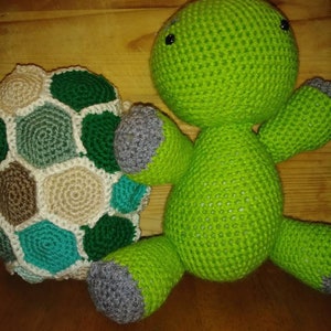 May include: A handmade green crochet turtle with gray feet and a matching shell. The shell is made of various colors, including white, green, and tan. The turtle has black button eyes and is sitting on a wooden surface.