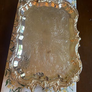 May include: A rectangular silver-toned serving tray featuring ornate, embossed scrollwork patterns across the centre surface. The tray has a raised, scalloped edge with intricate detailing and two decorative handles on opposite ends.