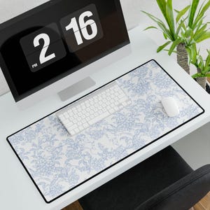 May include: A rectangular desk mat featuring a white background with a repeating light blue floral and botanical pattern. The mat has a thin black border and is positioned on a white desk surface beneath a computer keyboard and a white computer mouse.