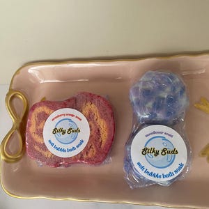 Puede incluir: Dos bombas de baño, una roja y naranja, la otra azul y morada, están sobre una bandeja rosa. Cada una está envuelta en plástico transparente y tiene una etiqueta circular que dice "Silky Suds soft bubble bath soak". La bomba de baño roja está etiquetada "strawberry orange scent".