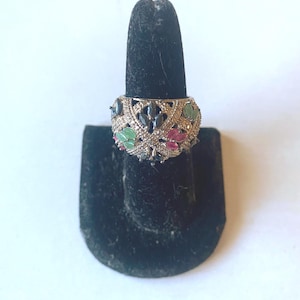 May include: A wide band ring featuring a lattice design encrusted with small clear stones. The ring is accented with marquise-cut gemstones in shades of green, pink, and dark blue, arranged in a repeating pattern across the metallic surface.