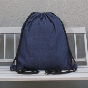 May include: A blue denim drawstring backpack with black drawstrings and trim. The bag is sitting on a white bench against a gray wall. The bag is made of a textured fabric.