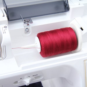 Pode incluir: Um close-up de uma m&aacute;quina de costura branca com um carretel de fio vermelho no enrola-bobina.