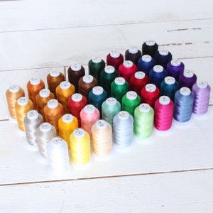 May include: A collection of 36 spools of embroidery thread in various colors, including red, blue, green, yellow, orange, brown, and white. The spools are arranged in a triangular shape on a white wooden surface.