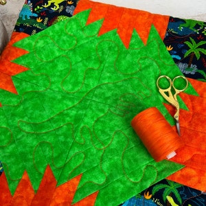 Pode incluir: Um quadrado de tecido verde com bordas de tecido laranja e preto. O tecido verde tem um desenho de espirais e la&ccedil;os costurados em vermelho. Uma bobina de fio laranja e um par de tesouras douradas est&atilde;o sobre o tecido.