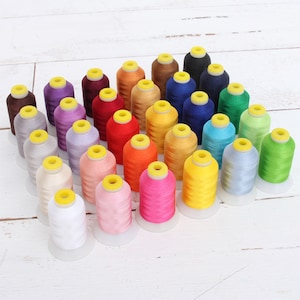May include: A collection of 30 spools of embroidery floss in various colors, including red, blue, green, yellow, orange, pink, purple, and white. The spools are arranged in a semi-circle on a white wooden surface.