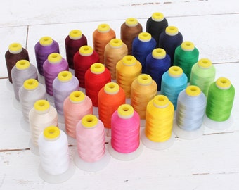 Puede incluir: Una colección de 30 carretes de hilo de bordar en varios colores, incluyendo rojo, azul, verde, amarillo, naranja, rosa, morado y blanco. Los carretes están dispuestos en un semicírculo sobre una superficie de madera blanca.