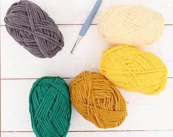 May include: Overhead shot of five skeins of yarn in various colors: gray, cream, yellow, green, and mustard. A blue crochet hook is also present. The yarn is arranged on a white wooden surface, ideal for crafting projects.