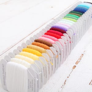May include: A collection of Sashiko thread spools arranged in a diagonal line, featuring a gradient of colors from white and yellow to brown, pink, red, purple, green, and blue. Each spool is labeled with 100% cotton, size 20/6, and made in Japan.