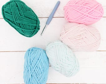 May include: An overhead shot of five yarn skeins in shades of green, pink, and blue, along with a crochet hook. The yarn is arranged on a white wooden surface. The colors are vibrant and the texture of the yarn is visible.