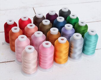 May include: A collection of twenty spools of embroidery thread arranged in a grid. The spools feature a variety of colors including shades of red, pink, orange, yellow, green, blue, purple, brown, gray, and black, each with a white plastic core.