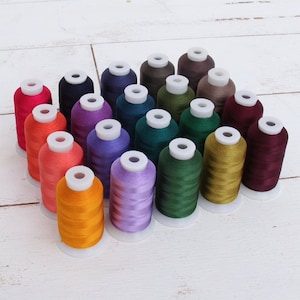 May include: A collection of 20 spools of embroidery thread in various colors, including red, orange, yellow, green, blue, purple, brown, and burgundy. The spools are arranged in a triangular shape on a white wooden surface.