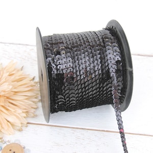May include: A spool of black sequin trim. The trim is made of small, round sequins that are sewn together in a continuous line. The spool is made of black plastic and has a white label.