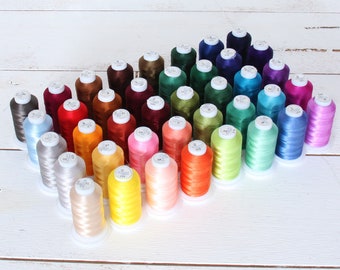May include: A collection of 36 spools of embroidery thread in various colors, arranged in a heart shape on a white wooden surface.