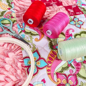Pode incluir: Um close-up de um projeto de acolchoado com retalhos de tecido coloridos e carret&eacute;is de fio vermelho, rosa e verde. Tamb&eacute;m &eacute; poss&iacute;vel ver um par de tesouras e um aro de bordado.