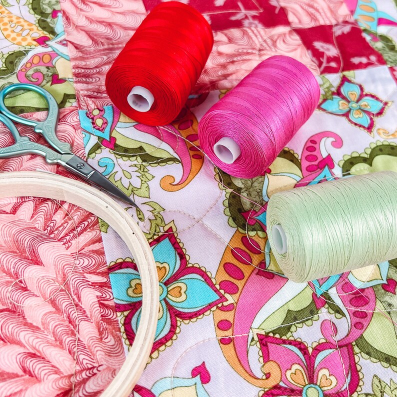 Pode incluir: Um close-up de um projeto de acolchoado com retalhos de tecido coloridos e carret&eacute;is de fio vermelho, rosa e verde. Tamb&eacute;m &eacute; poss&iacute;vel ver um par de tesouras e um aro de bordado.