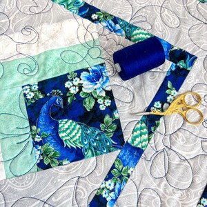 Pode incluir: Um close-up de um edredom com um design de pav&atilde;o. O edredom &eacute; feito de diferentes tecidos, incluindo uma estampa floral azul e verde, um tecido verde claro e um tecido branco com um padr&atilde;o paisley. H&aacute; um carretel de fio azul e um par de tesouras douradas no edredom.