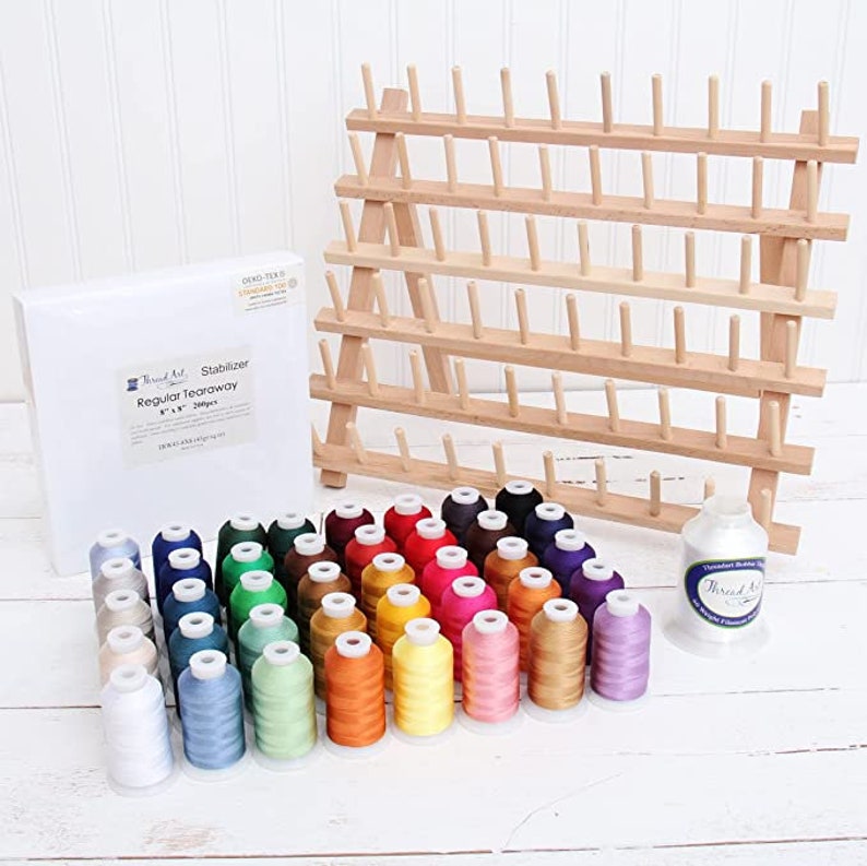 May include: A set of 48 spools of embroidery thread in various colors, including white, blue, green, red, orange, yellow, pink, purple, and brown. The spools are arranged in rows on a white surface. A wooden thread rack with 48 pegs is in the background. The box for the thread is labeled "Thread Art Stabilizer Regular Tearaway 8" x 8" 200 yds."