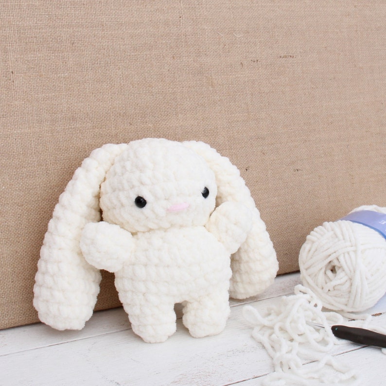 May include: A white crocheted bunny rabbit toy with long floppy ears. The bunny has black eyes and a pink nose.