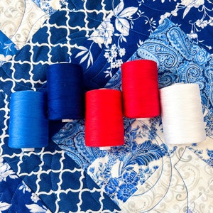 Pode incluir: Cinco carret&eacute;is de fio nas cores vermelho, branco e azul. Os carret&eacute;is est&atilde;o dispostos em uma fileira sobre um tecido estampado azul e branco.