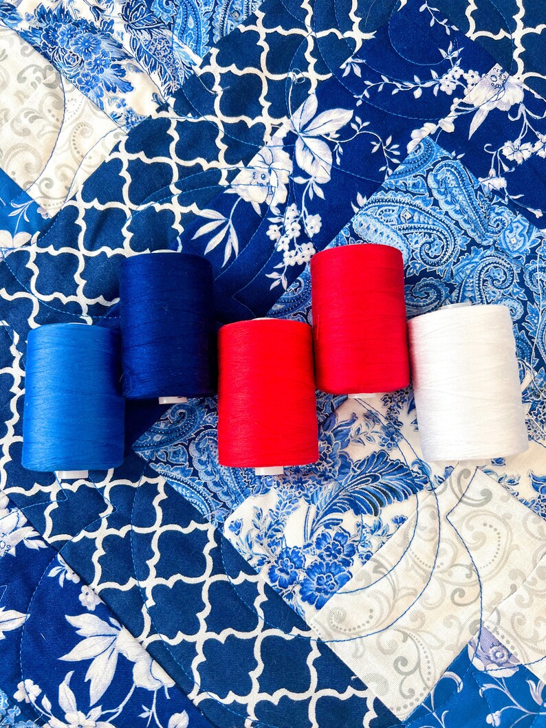 Pode incluir: Cinco carret&eacute;is de fio nas cores vermelho, branco e azul. Os carret&eacute;is est&atilde;o dispostos em uma fileira sobre um tecido estampado azul e branco.