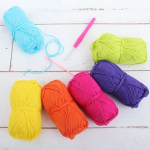 K&ouml;nnte beinhalten: Sechs Kn&auml;uel Wolle in verschiedenen Farben: Gelb, Orange, Rosa, Lila, Gr&uuml;n und Blau. Ein rosa H&auml;kelnadel liegt auf der wei&szlig;en Holzoberfl&auml;che.