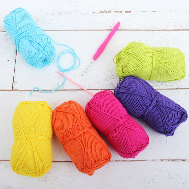 K&ouml;nnte beinhalten: Sechs Kn&auml;uel Wolle in verschiedenen Farben: Gelb, Orange, Rosa, Lila, Gr&uuml;n und Blau. Ein rosa H&auml;kelnadel liegt auf der wei&szlig;en Holzoberfl&auml;che.
