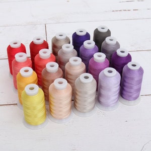 May include: A collection of 20 spools of embroidery thread in various colors, including yellow, orange, red, pink, purple, blue, and gray. The spools are arranged in a semi-circle on a white wooden surface.