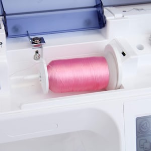 May include: A close-up of a pink thread spool in the bobbin winder of a white sewing machine.