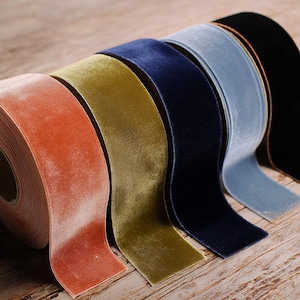 May include: A selection of velvet ribbon rolls in various colors, including coral, olive green, navy blue, and black. The ribbons are arranged side by side, showcasing their soft texture and color variations. The ribbons are on a wooden surface.