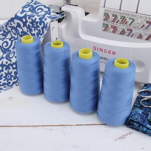 May include: Four spools of blue sewing thread with yellow tops, sitting on a white wooden surface. The spools are in front of a white sewing machine with the word "SINGER" printed on it.