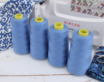 May include: Four spools of blue sewing thread with yellow tops, sitting on a white wooden surface. The spools are in front of a white sewing machine with the word "SINGER" printed on it.