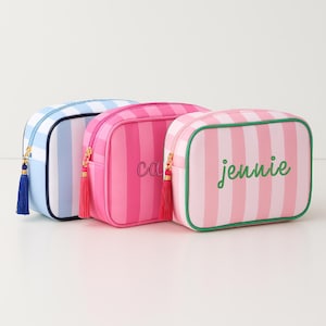 May include: Three striped cosmetic bags in blue, pink, and pink with green trim. The bags have gold zippers and matching tassels. The pink bag has the name "Carrie" embroidered on it, and the other pink bag has the name "jennie" embroidered on it.