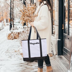 Może przedstawiać: Biała torba typu tote z czarnymi uchwytami i haftem „Magnolia” w kolorze czarnym na przodzie. Torbę trzyma osoba ubrana w biały sweter i dżinsy.